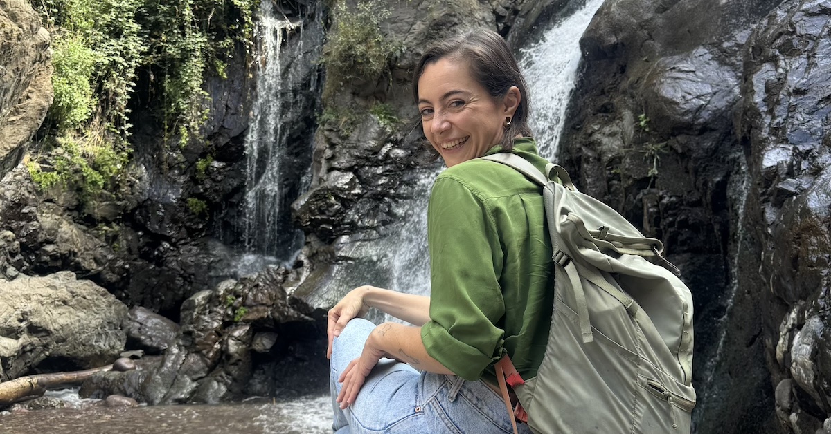 50AE6D8E-0F38-4FE6-997D-2E7A7C0DAAEE copy Frances sat down and smiling with a waterfall in the background.